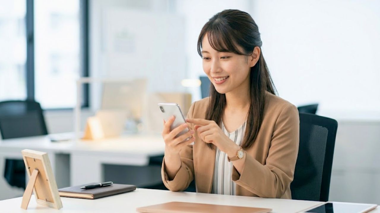 明るいオフィスのデスクで、スマートフォンをスマートに操作し、角を立てずに会社の飲み会への欠席を伝える出欠連絡を送るシングルマザー